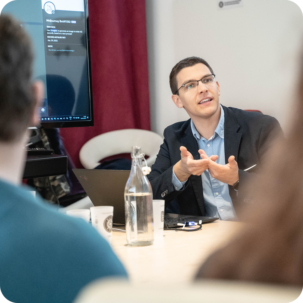 Lukas Kränkl sitzt an einem Tisch in einem Workshop und erklärt etwas